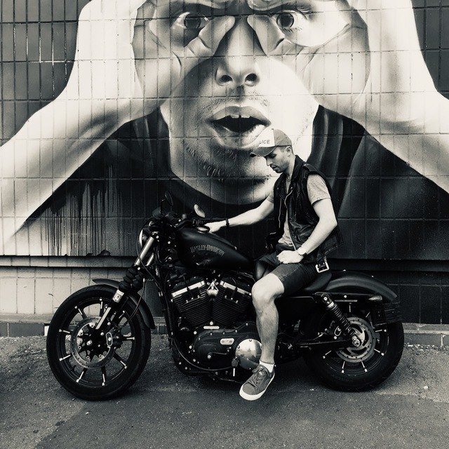 Black-and-white photo of Serhii Ksonzenko sitting on a black motorcycle in front of a large street mural of a man looking through his hands as if through binoculars