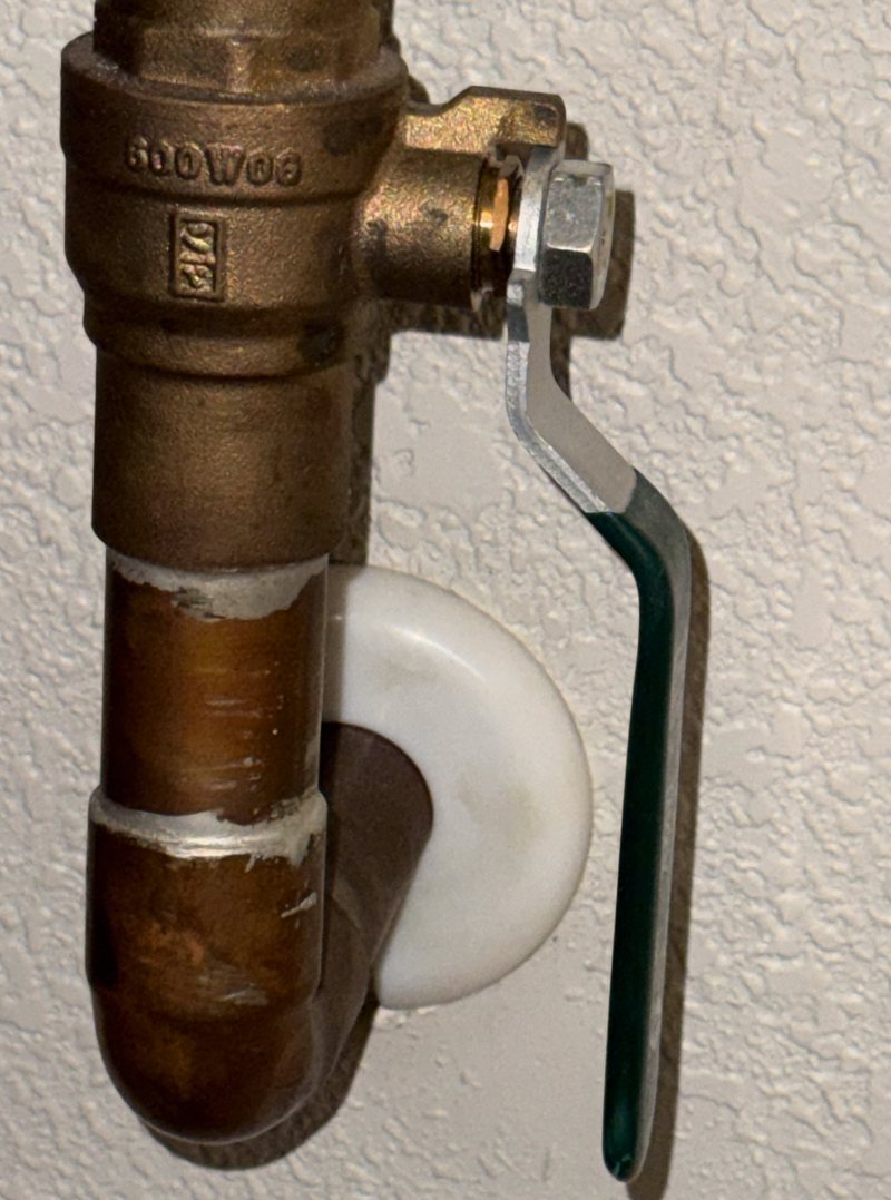 A brass ball valve with a long lever handle on a copper water pipe coming out of a wall; the handle runs along the pipe, which means the valve is open